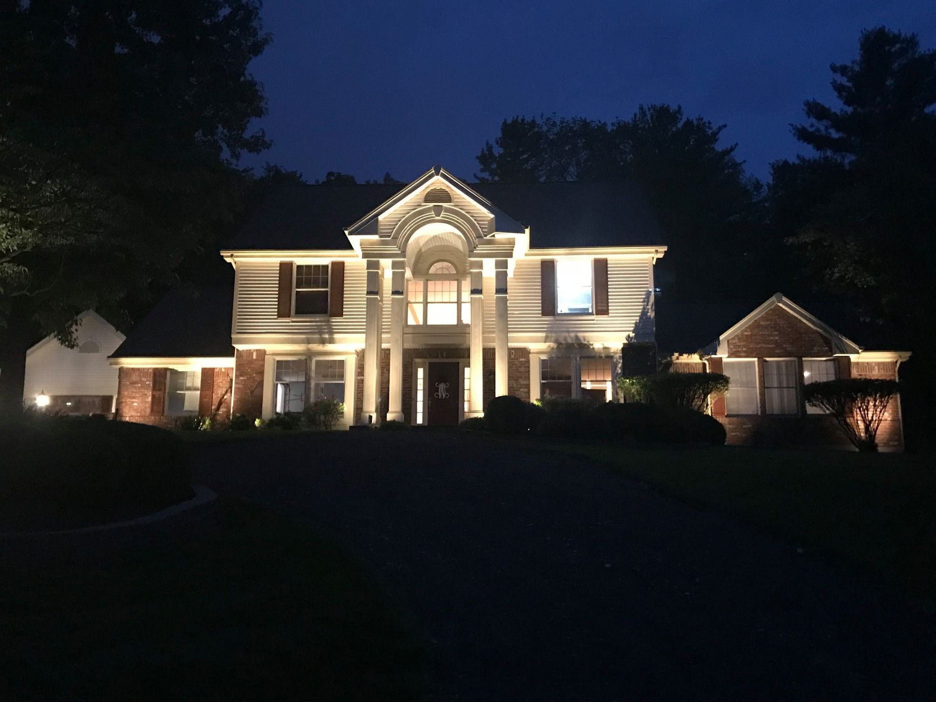 Illuminated suburban house at night with warm exterior lights and dark trees surrounding it