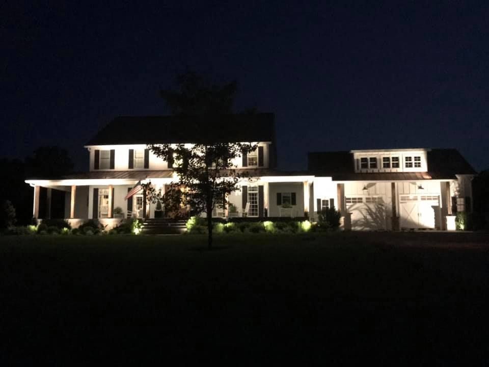 Lit house exterior at night with white facade, porch lights, and a tree in front