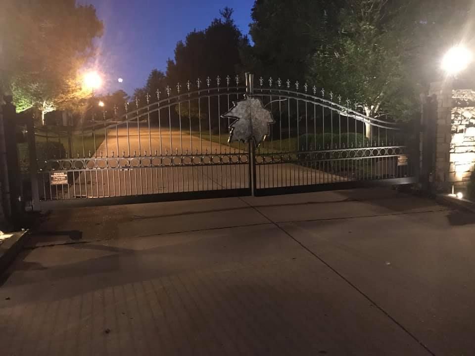 Closed wrought-iron gate across a driveway at night, lit by streetlights and flanked by trees.