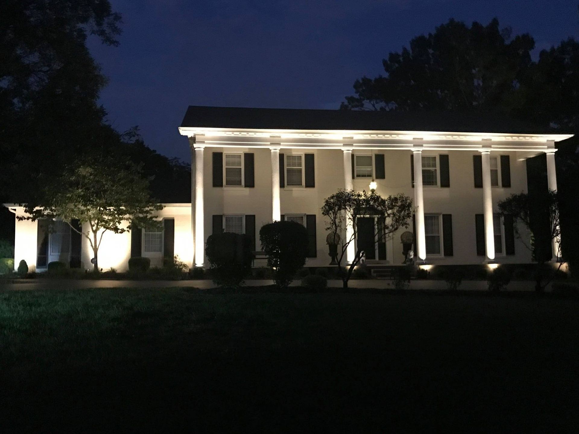 Illuminated white mansion at night with columns and lit windows, surrounded by dark trees and lawn