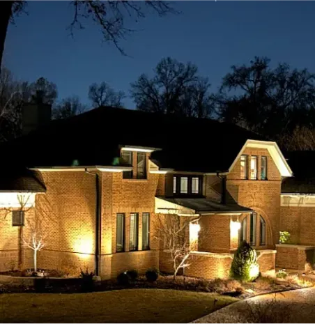 Illuminated brick house at night with warm exterior lights and dark trees in the background