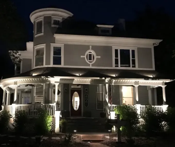 Victorian-style gray house with white trim, lit warmly at night, with a wraparound porch and tower.
