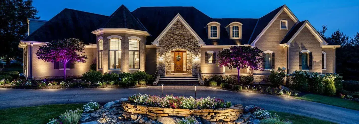 Large stone house at night with lit windows, purple-lit trees, and a landscaped front yard with a fountain