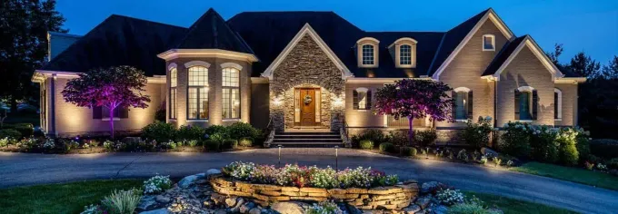 Large luxury house at night with lit windows, stone facade, and colorful garden landscape lighting