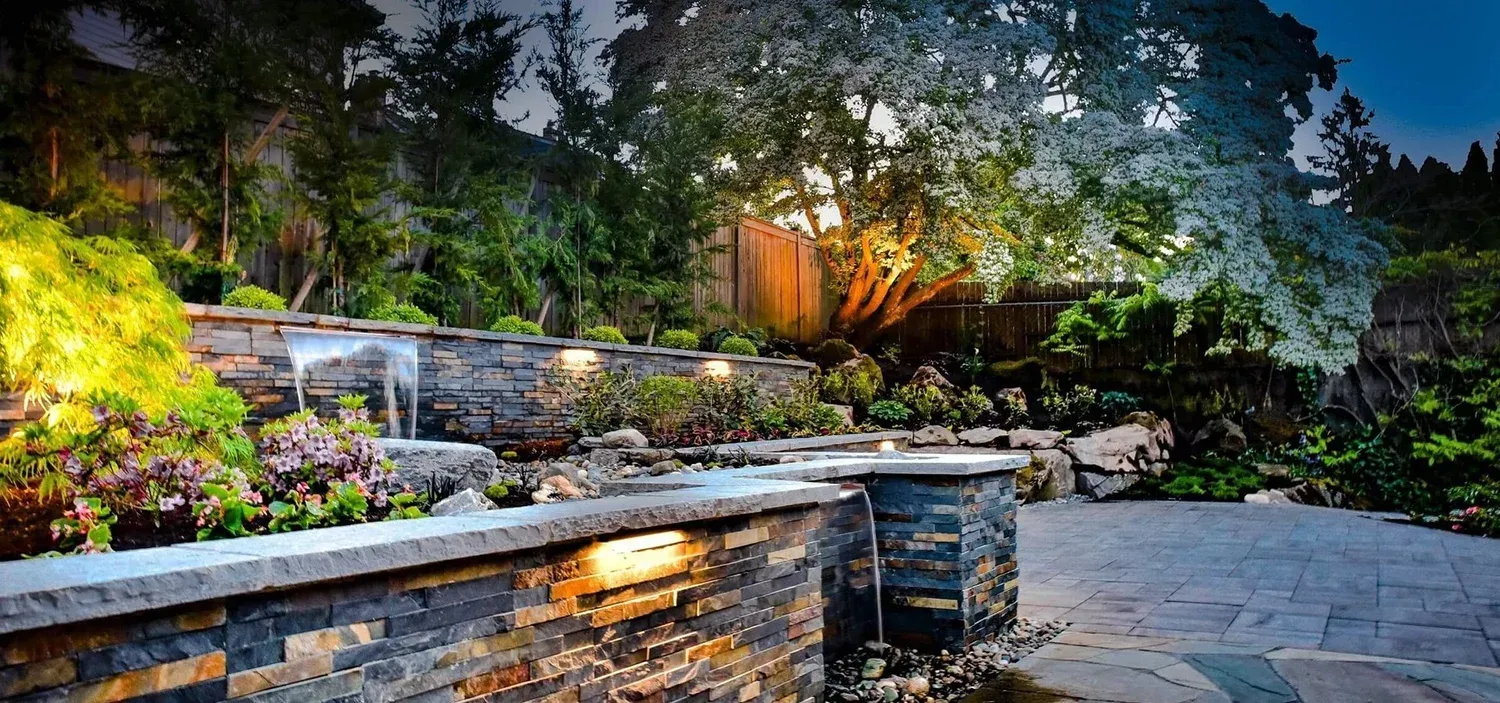 Stone patio and terraced garden with colorful flowers, lit steps, and trees at dusk