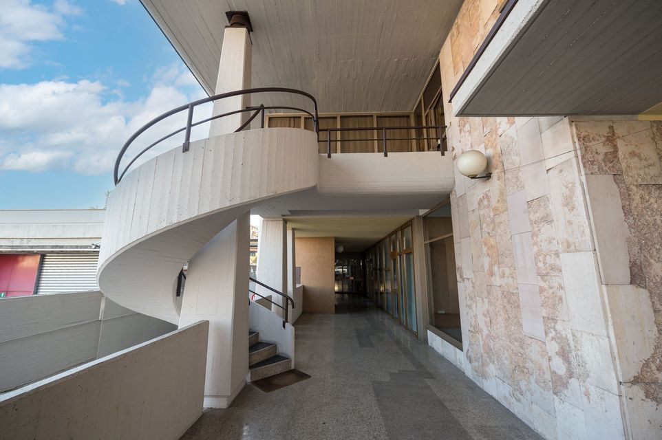 Vista esterna di un edificio con scala a chiocciola, passerella e muro in pietra usurata contro un cielo nuvoloso.
