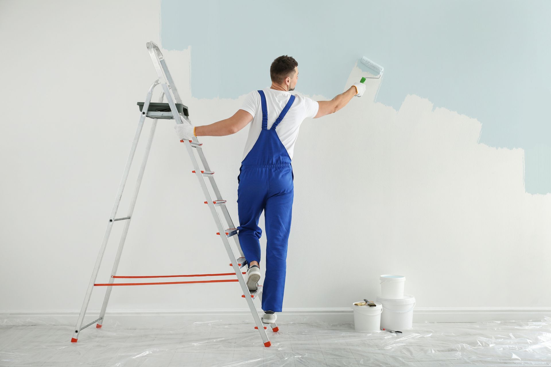 A painter in blue overalls paints a wall light blue with a roller while standing on a ladder.