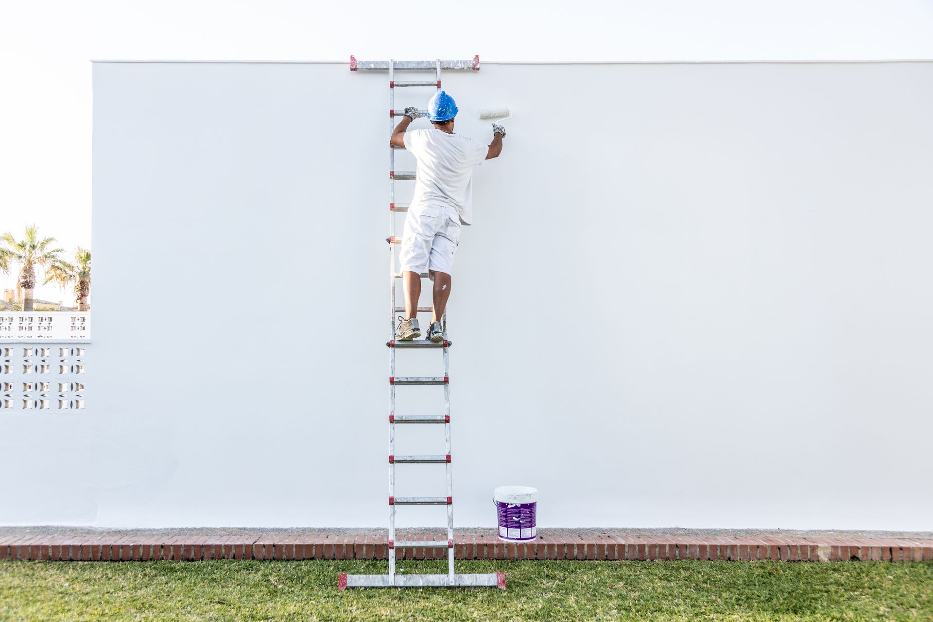 A painter is applying white paint to an exterior wall, with a paint bucket below. A painter is applying white paint to an exterior wall, with a paint bucket below.