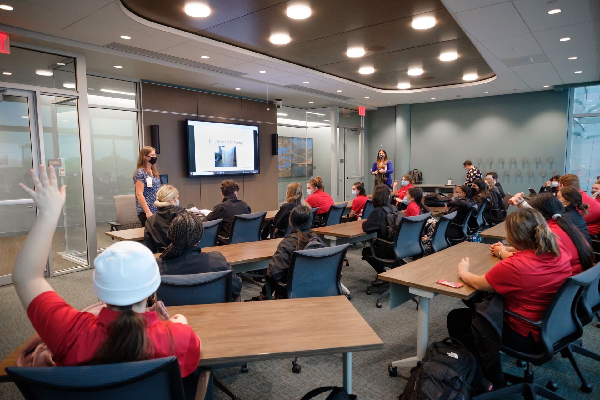 Classroom scene: students in red shirts listen to a presentation. One student raises their hand.