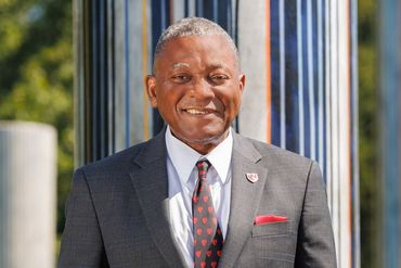 Smiling man in a suit, red tie, and pocket square, in front of a striped pillar outdoors.