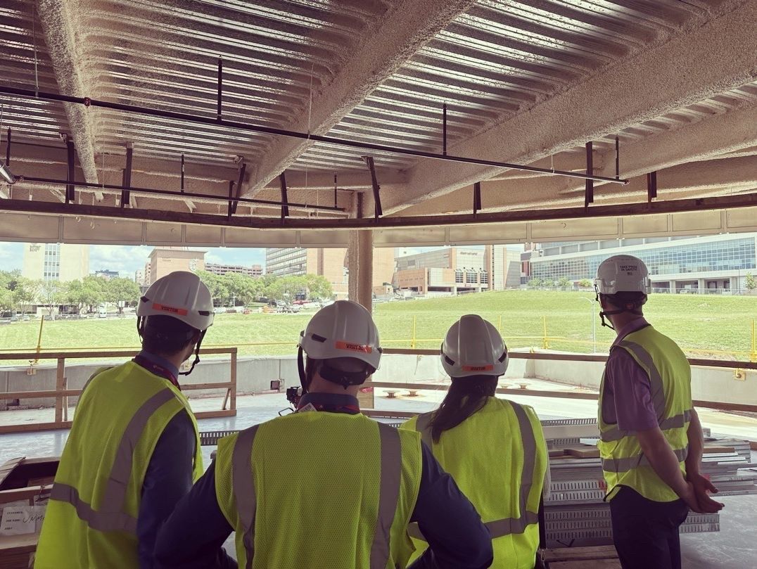 Four people in hard hats and vests, viewing construction site. Outside, stadium and park.