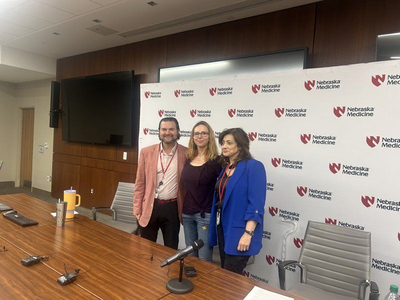 Three people stand in front of a Nebraska Medicine backdrop. Two women and a man pose near a table.