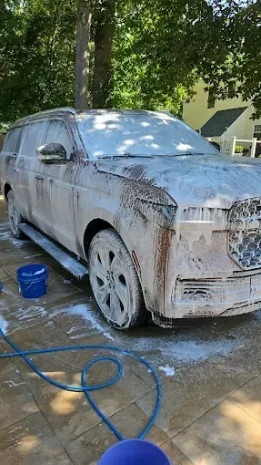 SUV covered in white foam, being washed outdoors with a hose and bucket.