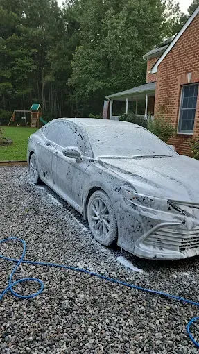A silver car covered in white foam suds, being washed on a gravel driveway next to a brick house.
