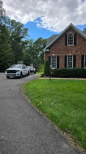White truck pulling trailer on driveway in front of brick house with black shutters, green lawn, and blue sky.