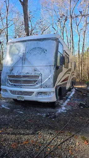 Motorhome covered in white foam, being washed outdoors, surrounded by trees.