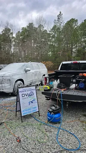 Car covered in foam, next to a truck bed with detailing equipment. Blue air compressor, sign in front, wooded background.
