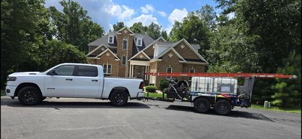 White pickup truck towing a trailer with equipment parked in front of a brick house.