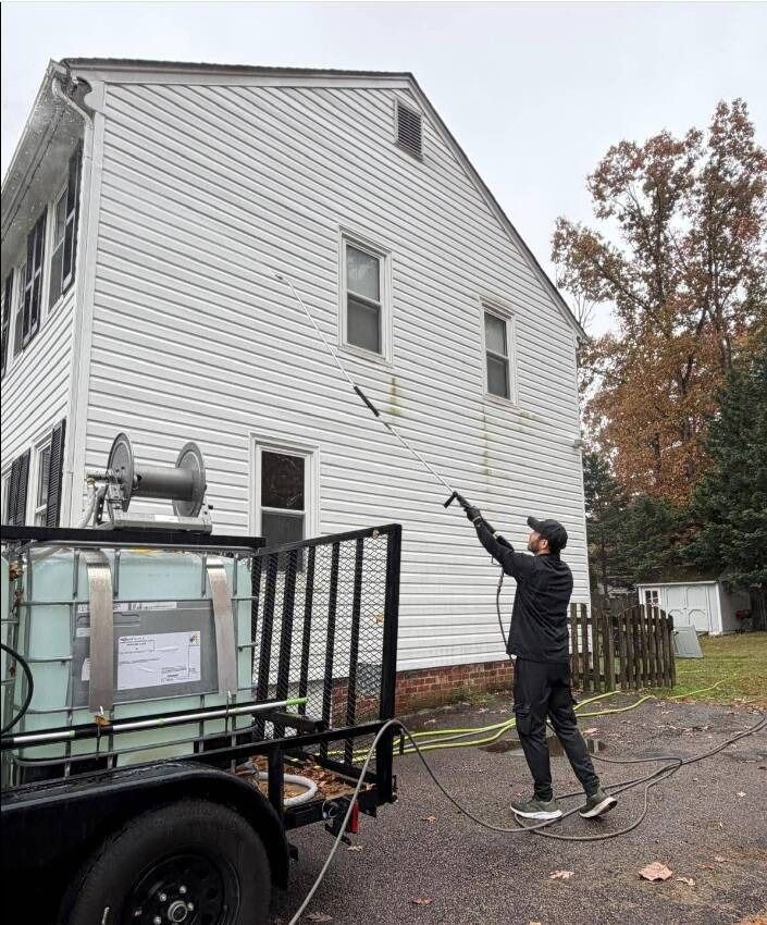 A person power washing a white house's siding. A trailer with equipment is visible.