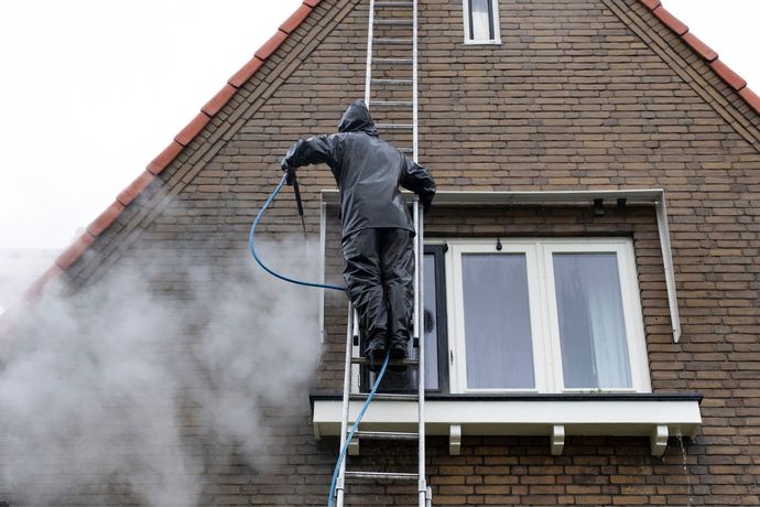 Person in rain gear on ladder power washing a brick house, creating steam.