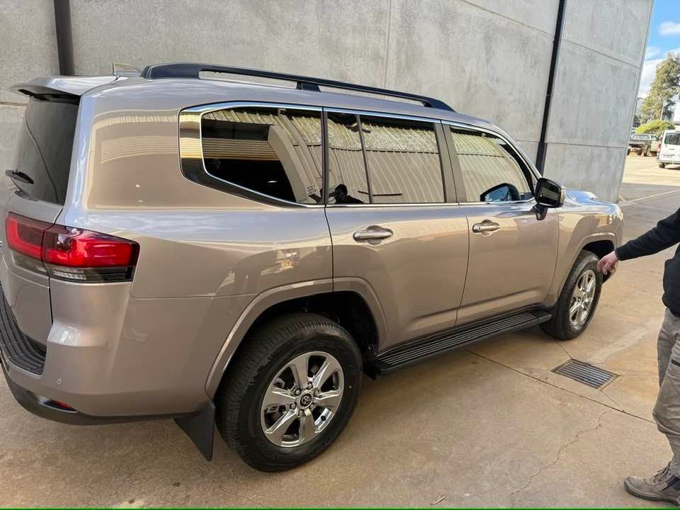 Tan Toyota Land Cruiser parked next to a gray wall; black roof rack, chrome wheels.