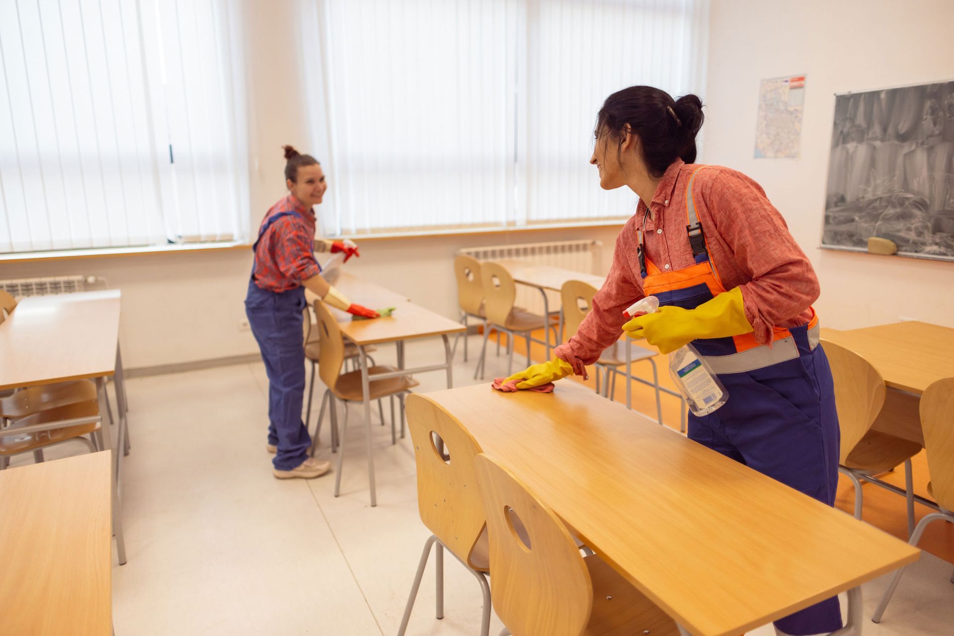 Two women are cleaning tables and chairs in a classroom.