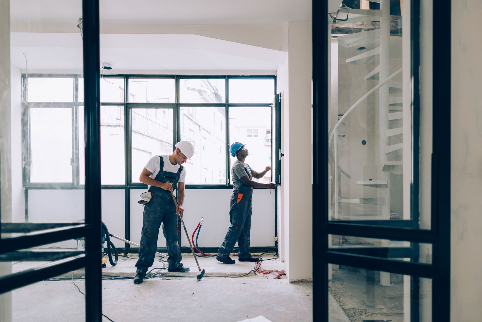 Two construction workers are working in an empty room with a spiral staircase in the background.