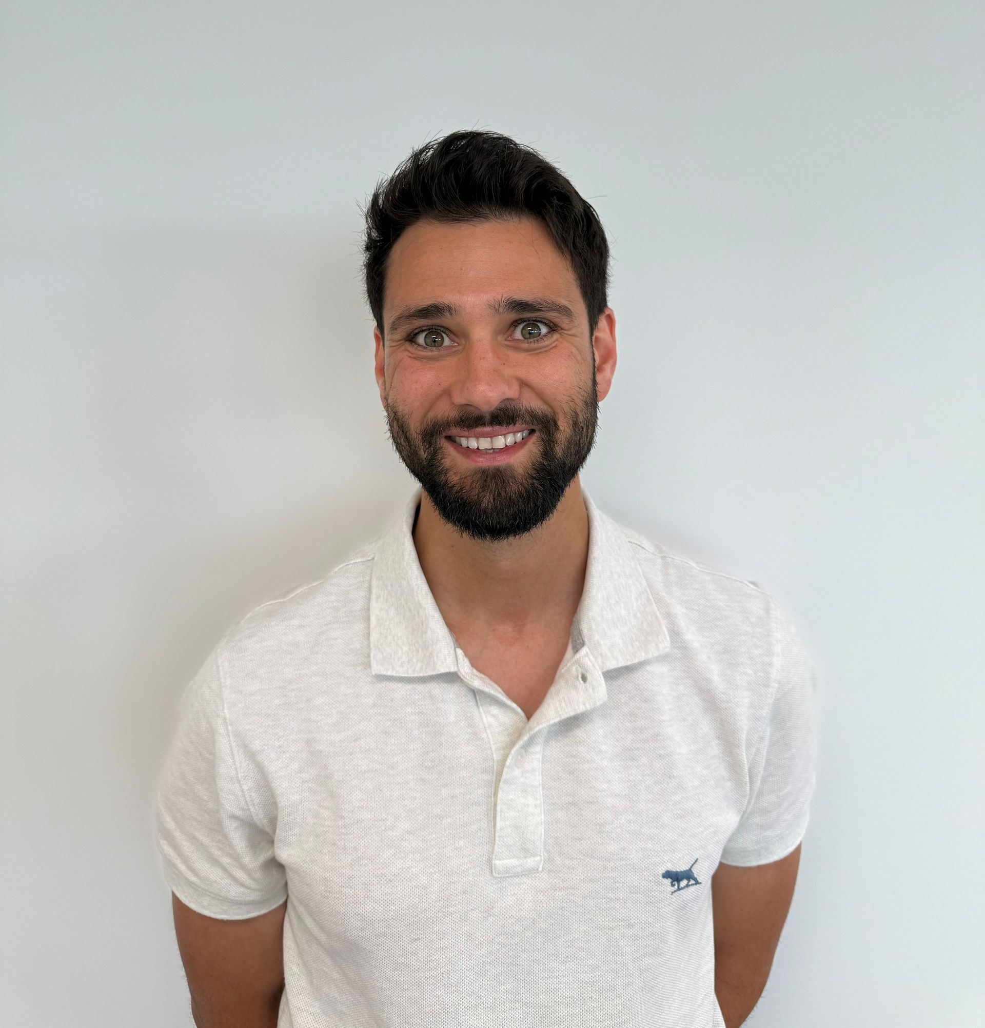 A man with a beard is wearing a white polo shirt and smiling.