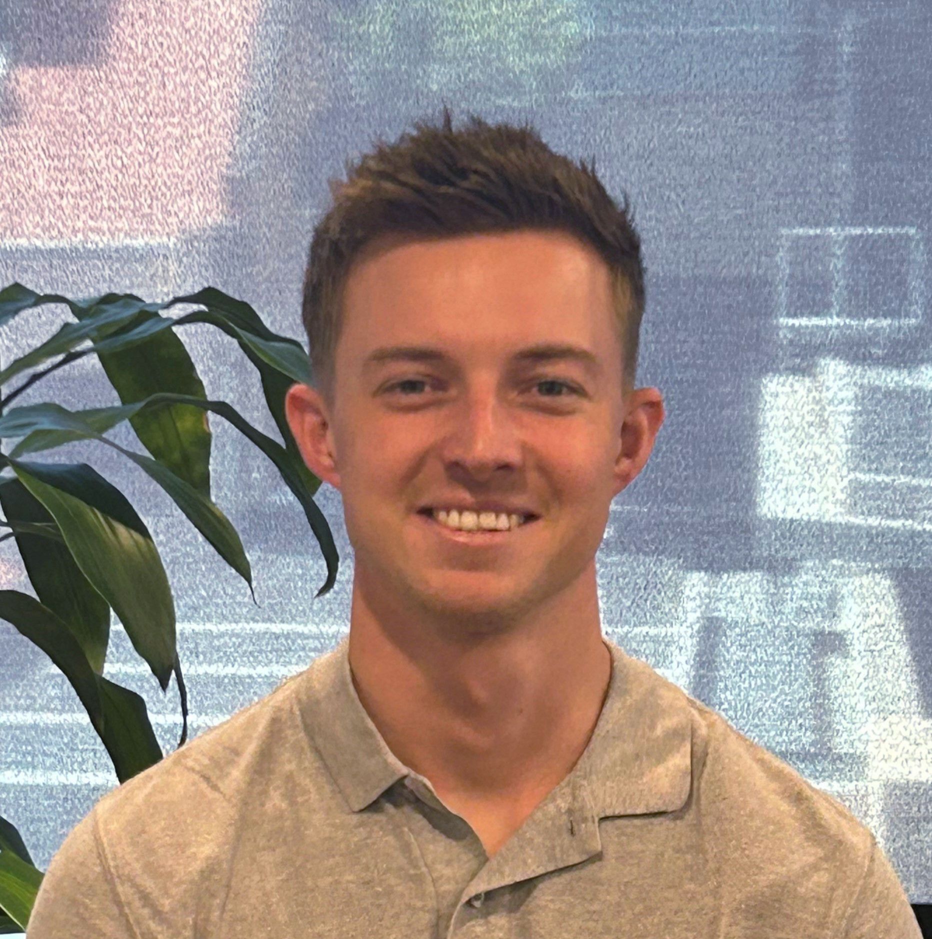 A young man in a grey shirt is smiling in front of a plant.