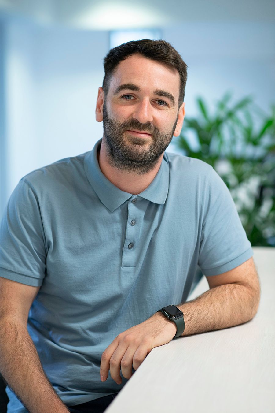 A man with a beard is sitting at a table wearing a blue polo shirt and a watch.