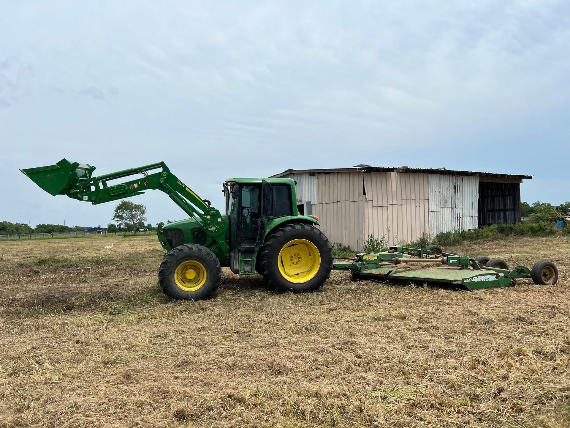 Green tractor with front loader parked in a dry field beside a small shed, towing farm equipment.