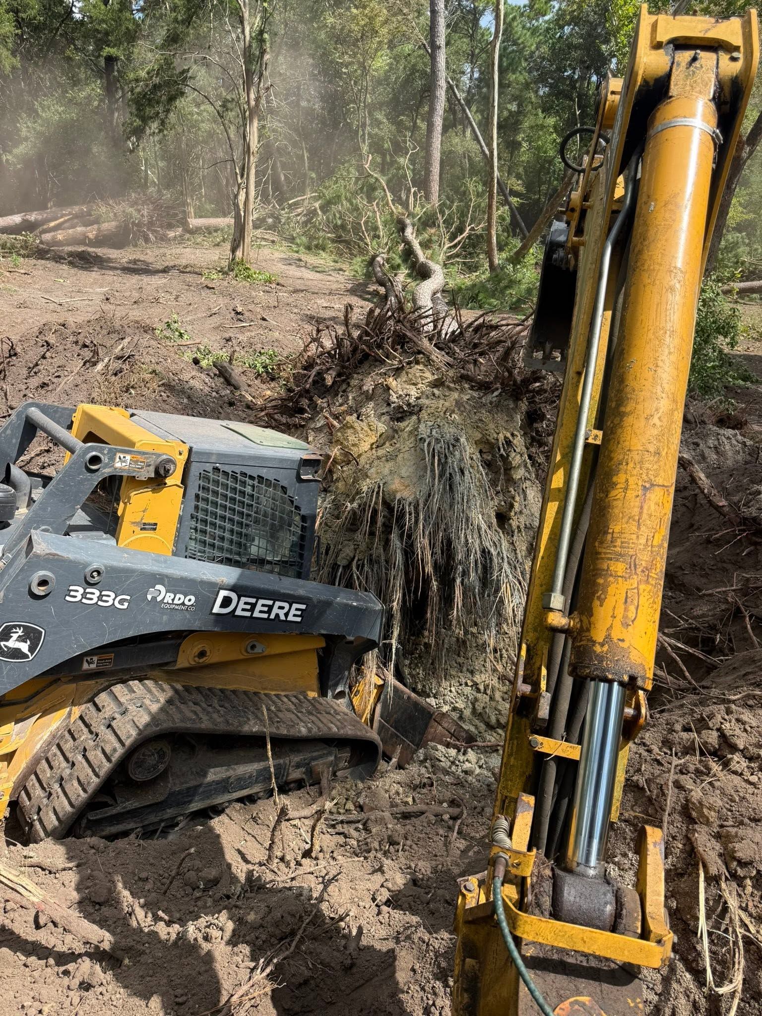 Yellow excavator bucket digging muddy ground beside a John Deere tracked machine in a wooded area