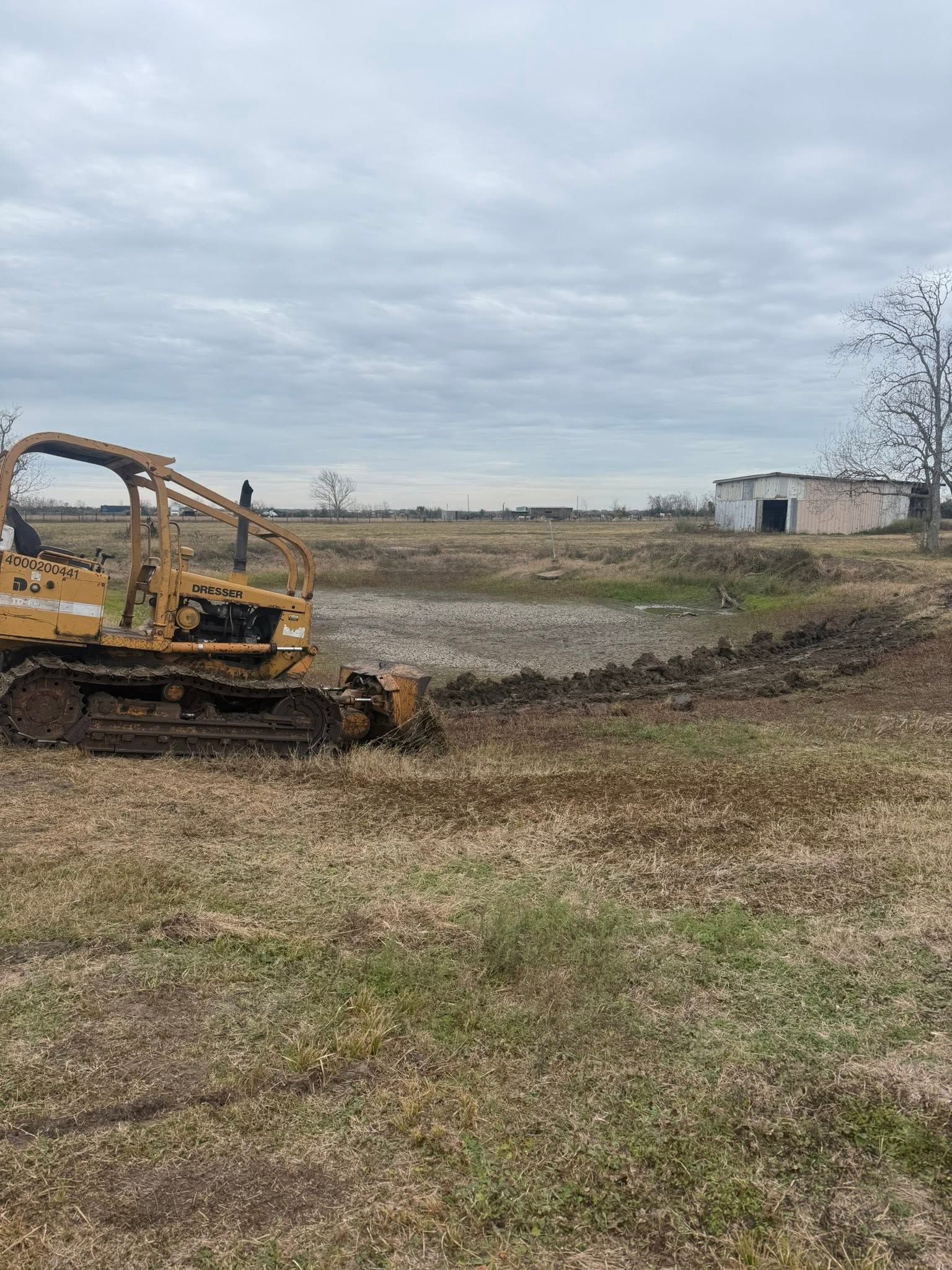Yellow bulldozer grading a muddy field under a cloudy sky with a small building in the distance