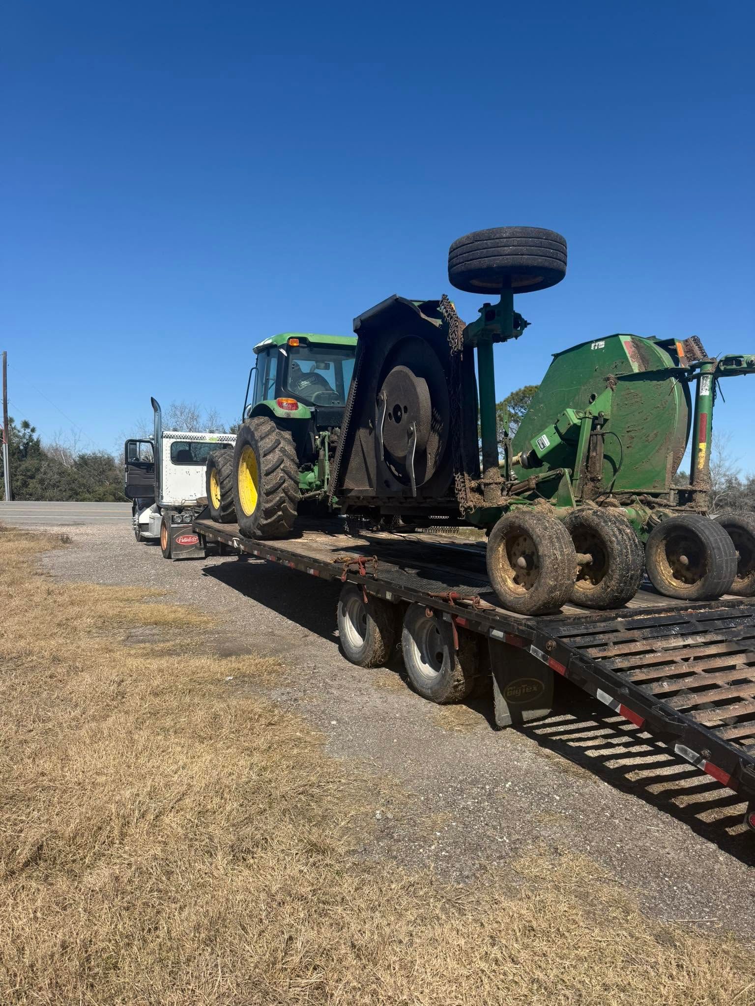 Green tractor on a flatbed trailer in a gravel lot under a clear blue sky.