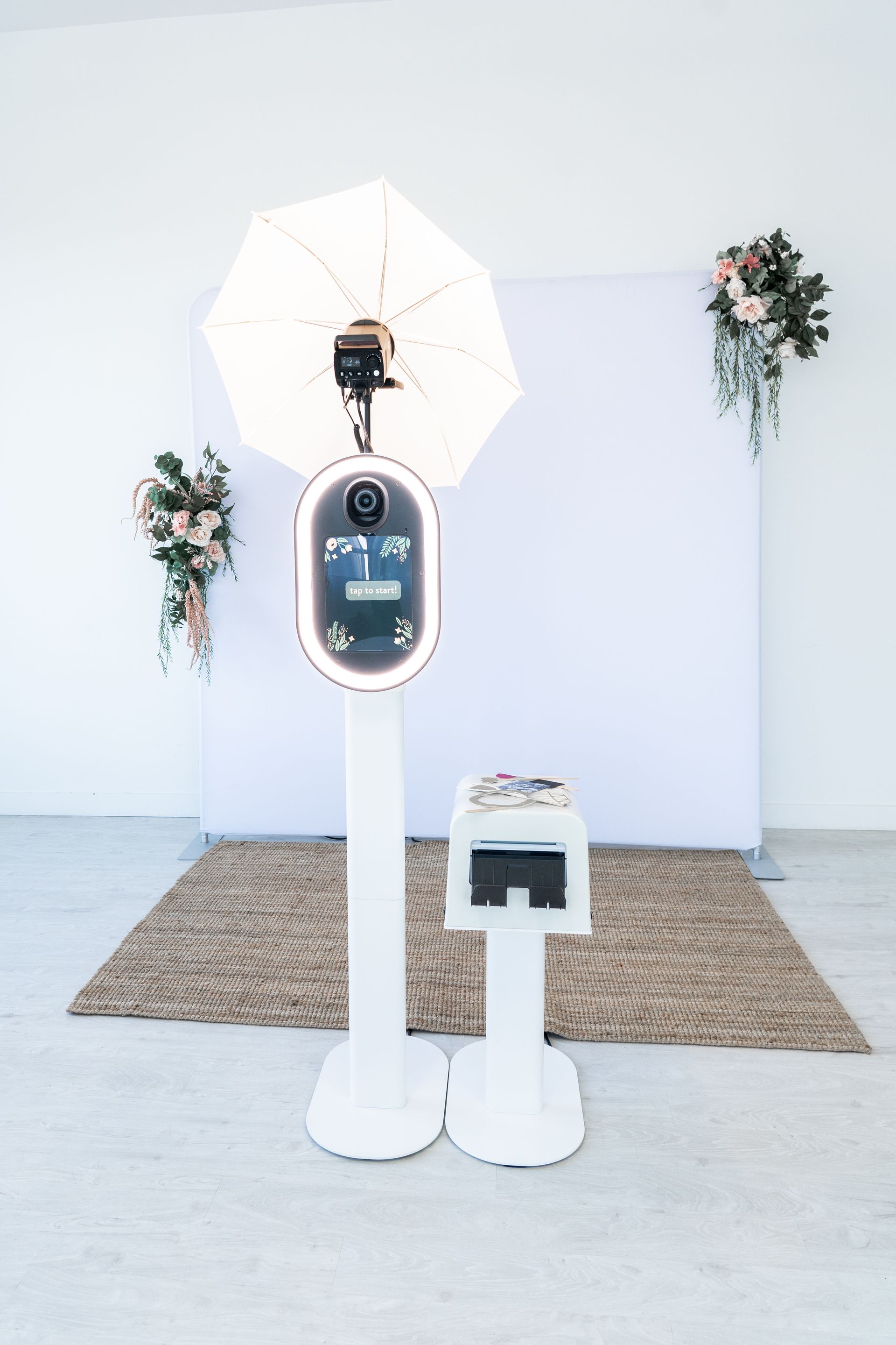 Photo booth setup with white backdrop, floral accents, and rug.