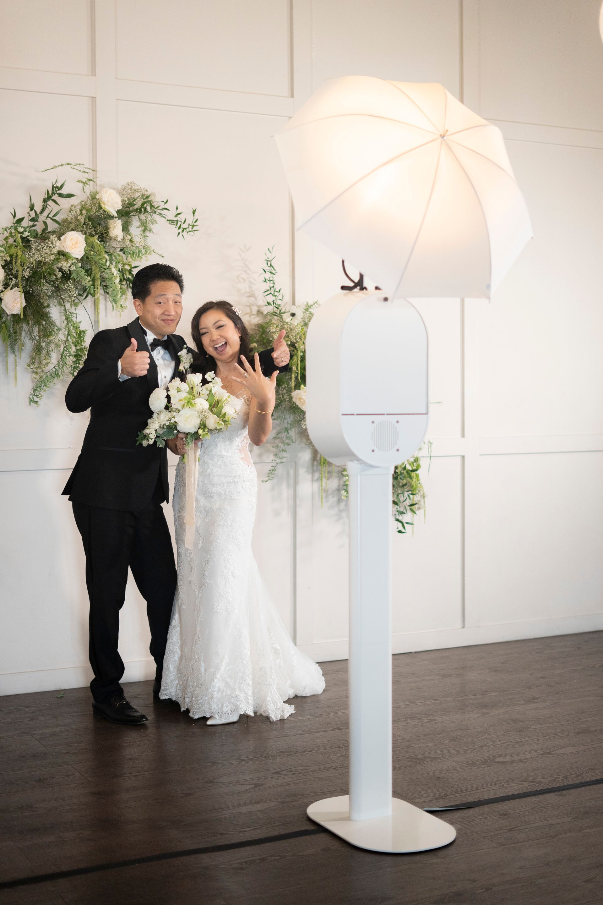 Newlyweds pose with thumbs up next to a white photo booth at a wedding.