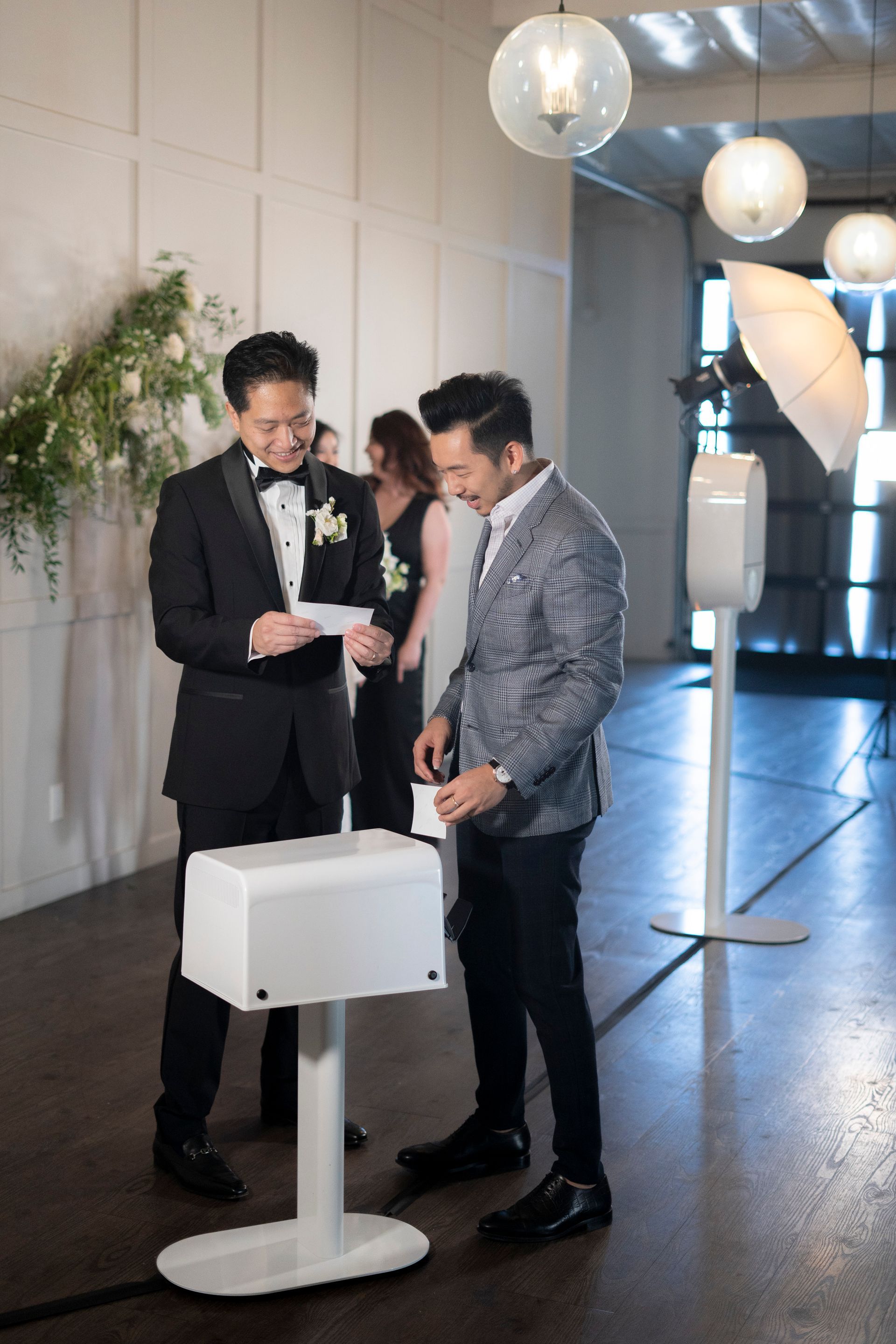 Two men in formal attire at a wedding, one reading a card, the other putting a card in a white box.