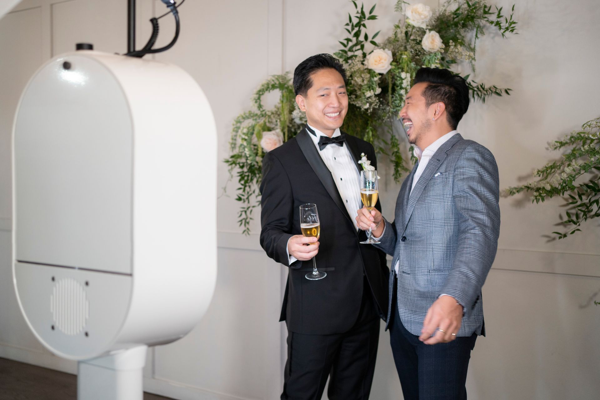Two men in formal attire, holding champagne flutes, smile by a photo booth at an event.