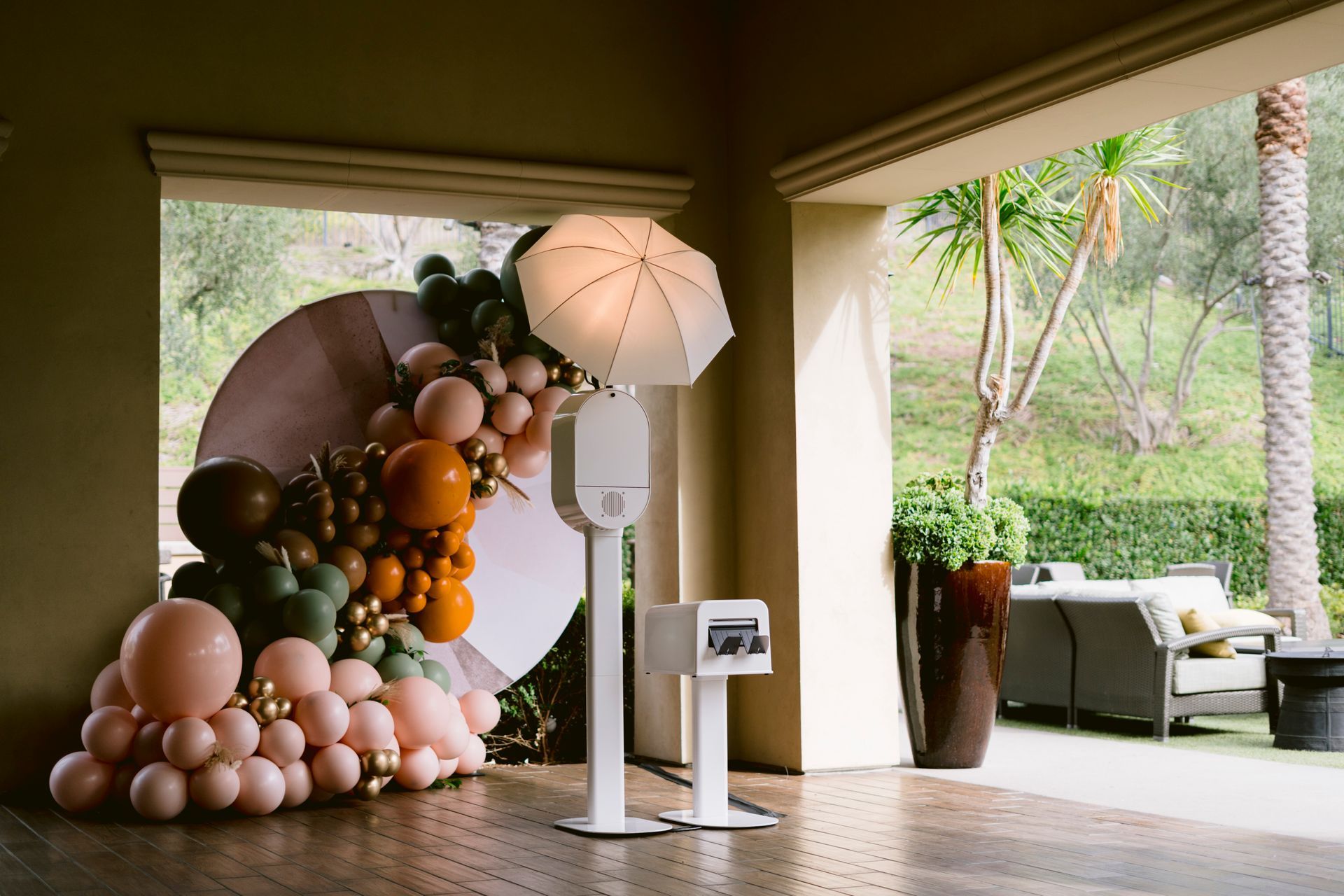Photo booth with balloons against an outdoor backdrop. White equipment, orange, green, and pink balloons.