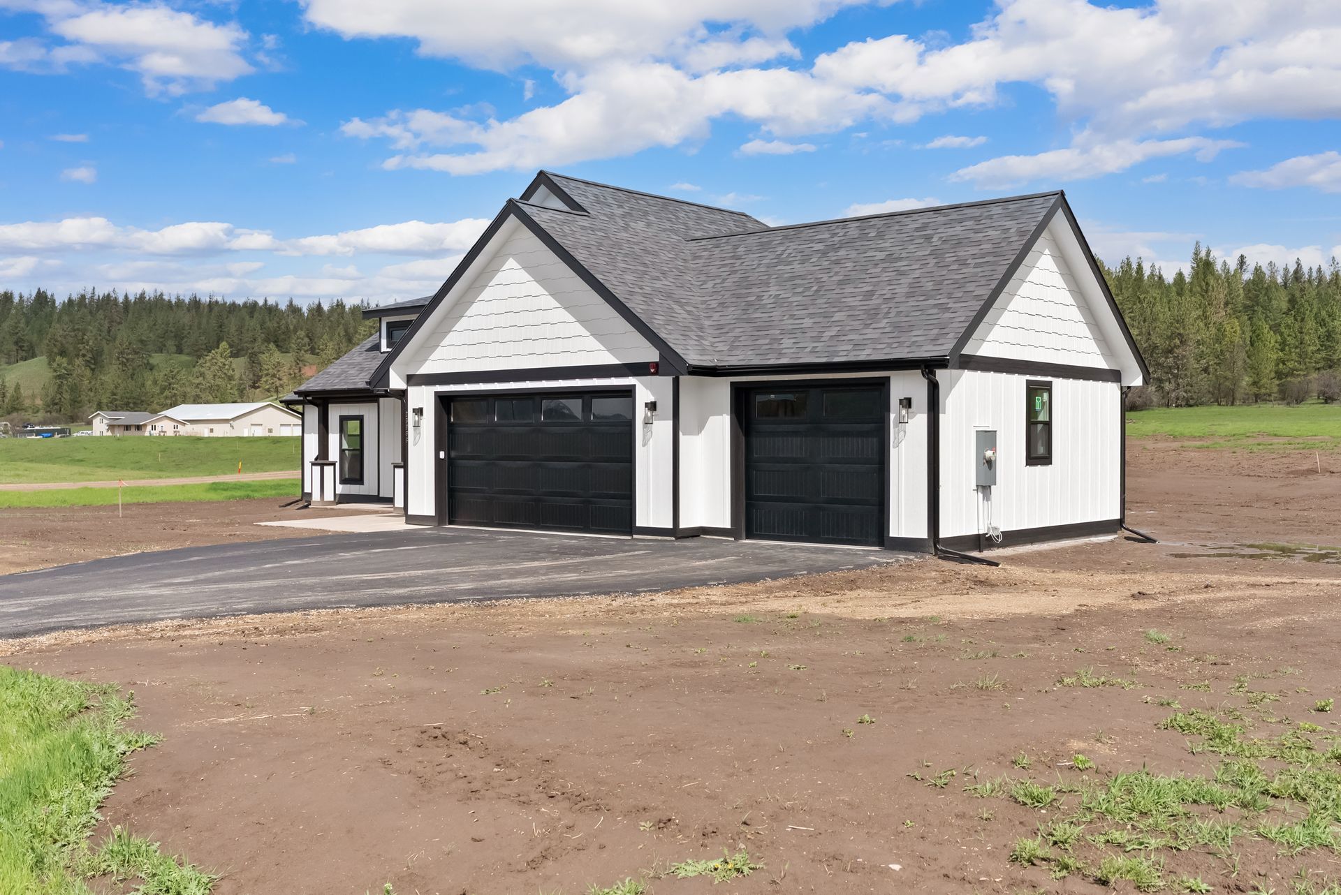 Small white house with dark roof and two garage doors on a dirt lot, surrounded by open field and trees