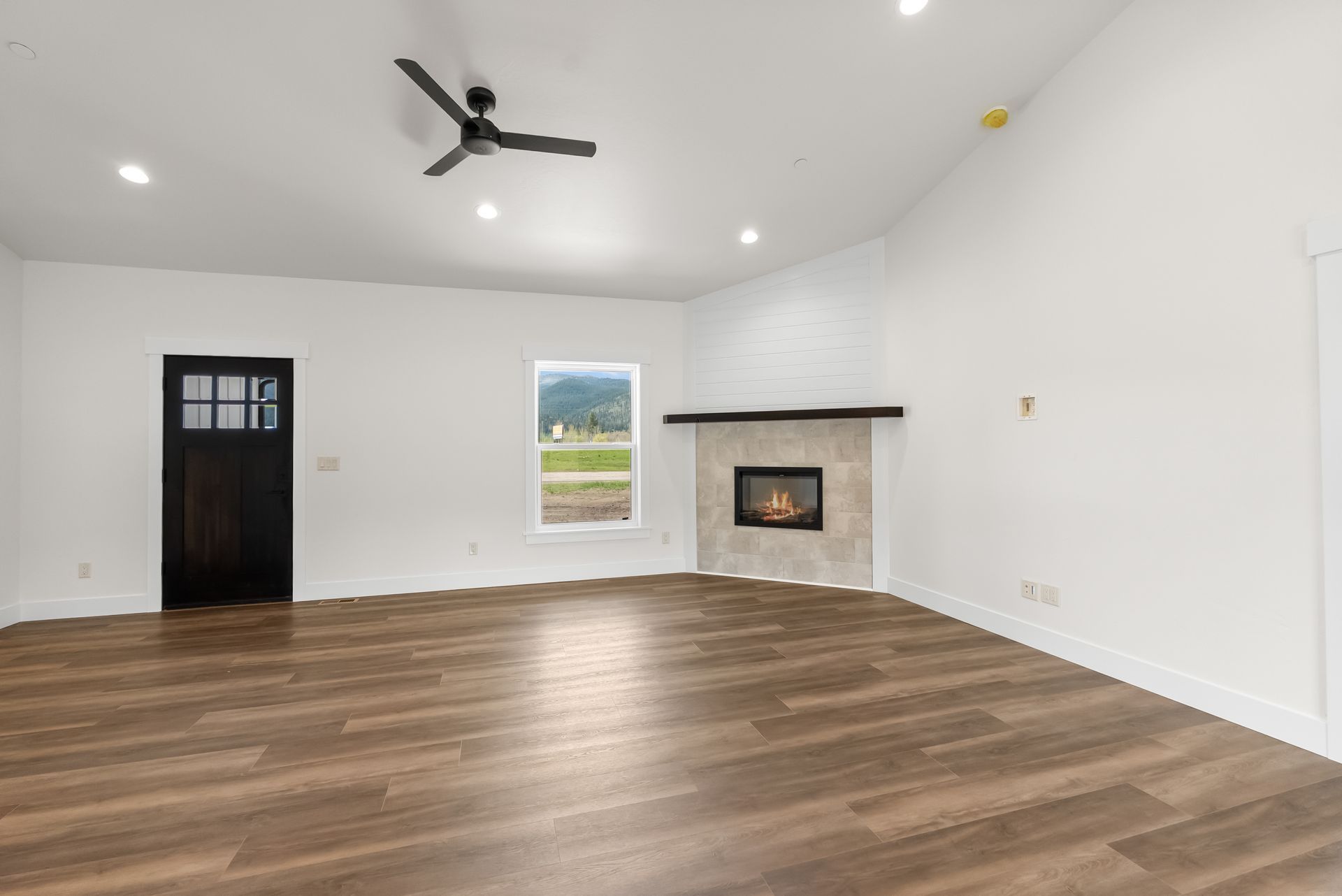 Empty white living room with wood floors, black ceiling fan, fireplace, and a window.