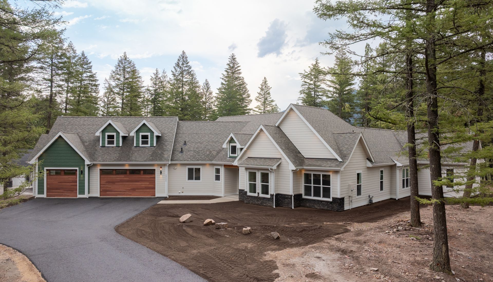 Large gray house with green roof and red garage in a wooded setting under cloudy skies