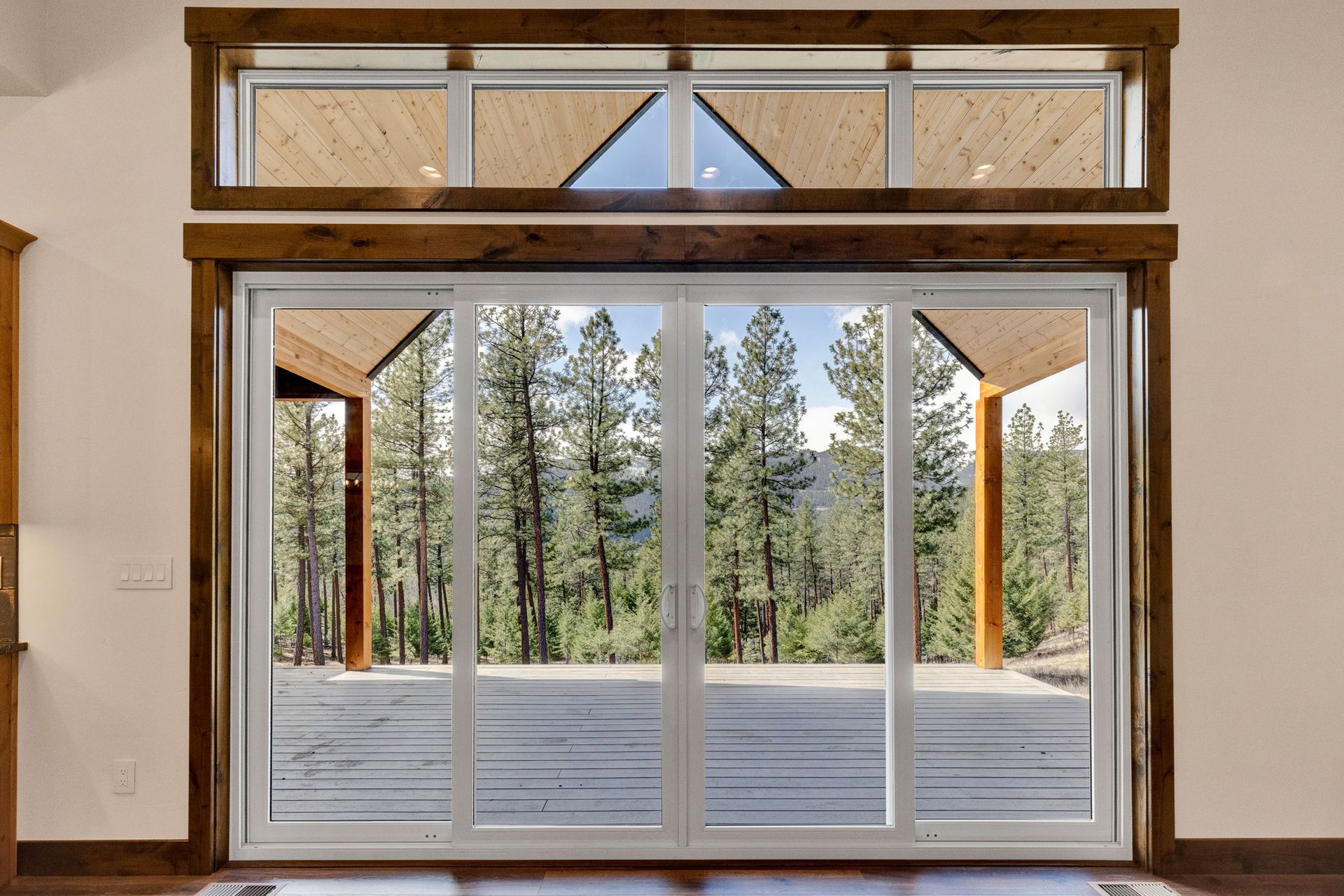 Large sliding glass doors opening to a wooden deck and pine trees outside