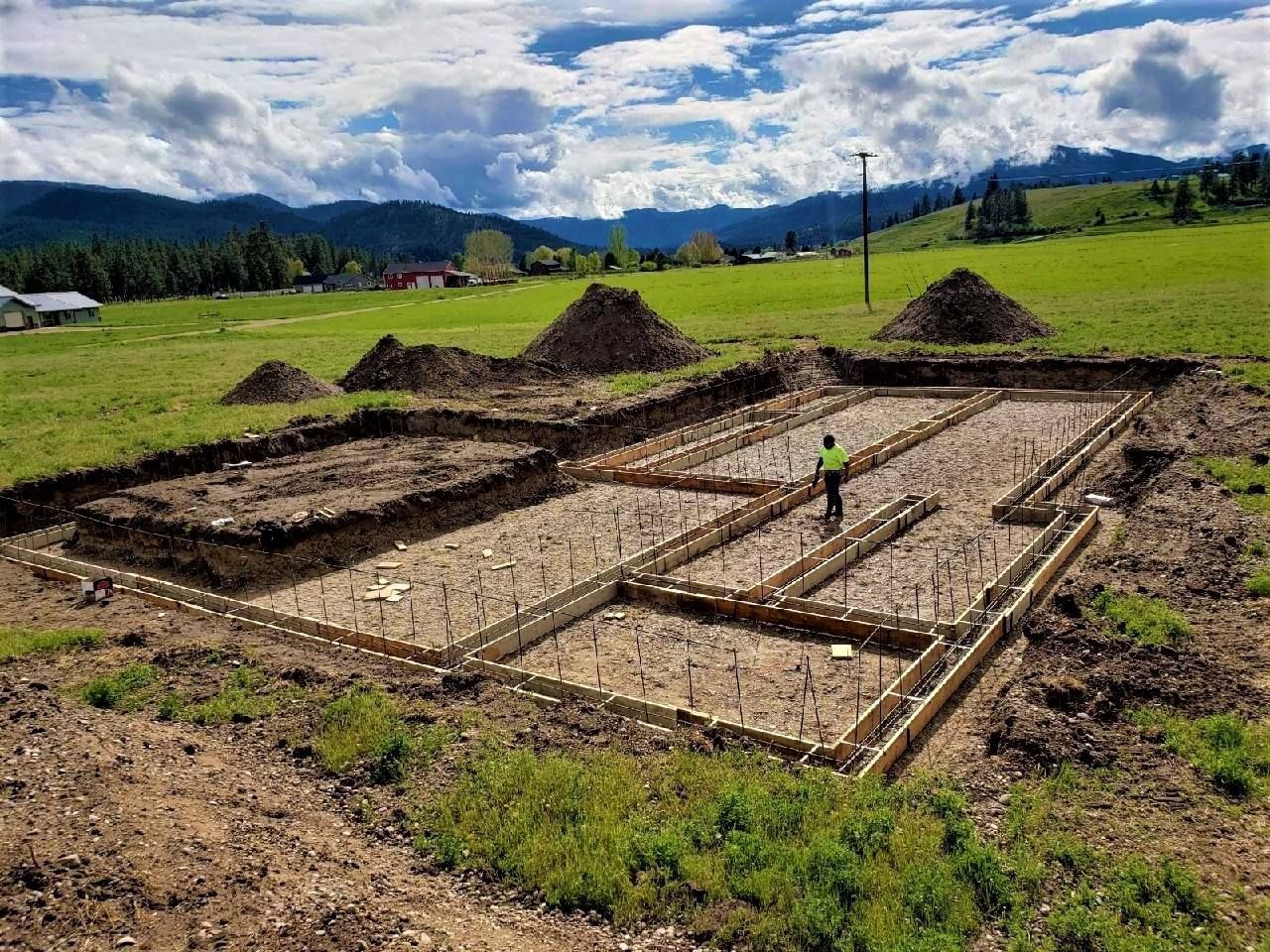 Foundation trenches at a rural construction site, with a worker in a green vest near dirt mounds and grassy fields.