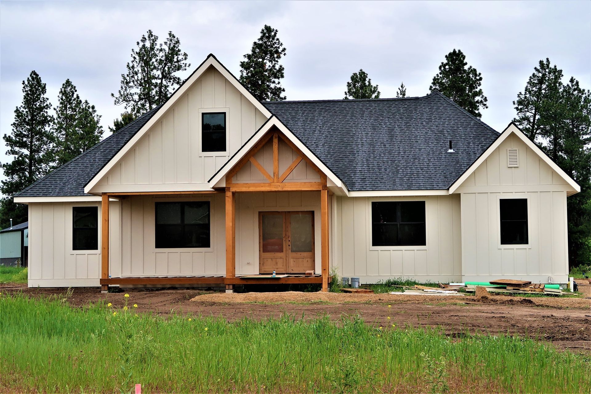 New cream-colored house with dark roof and front porch under construction, surrounded by grass and trees