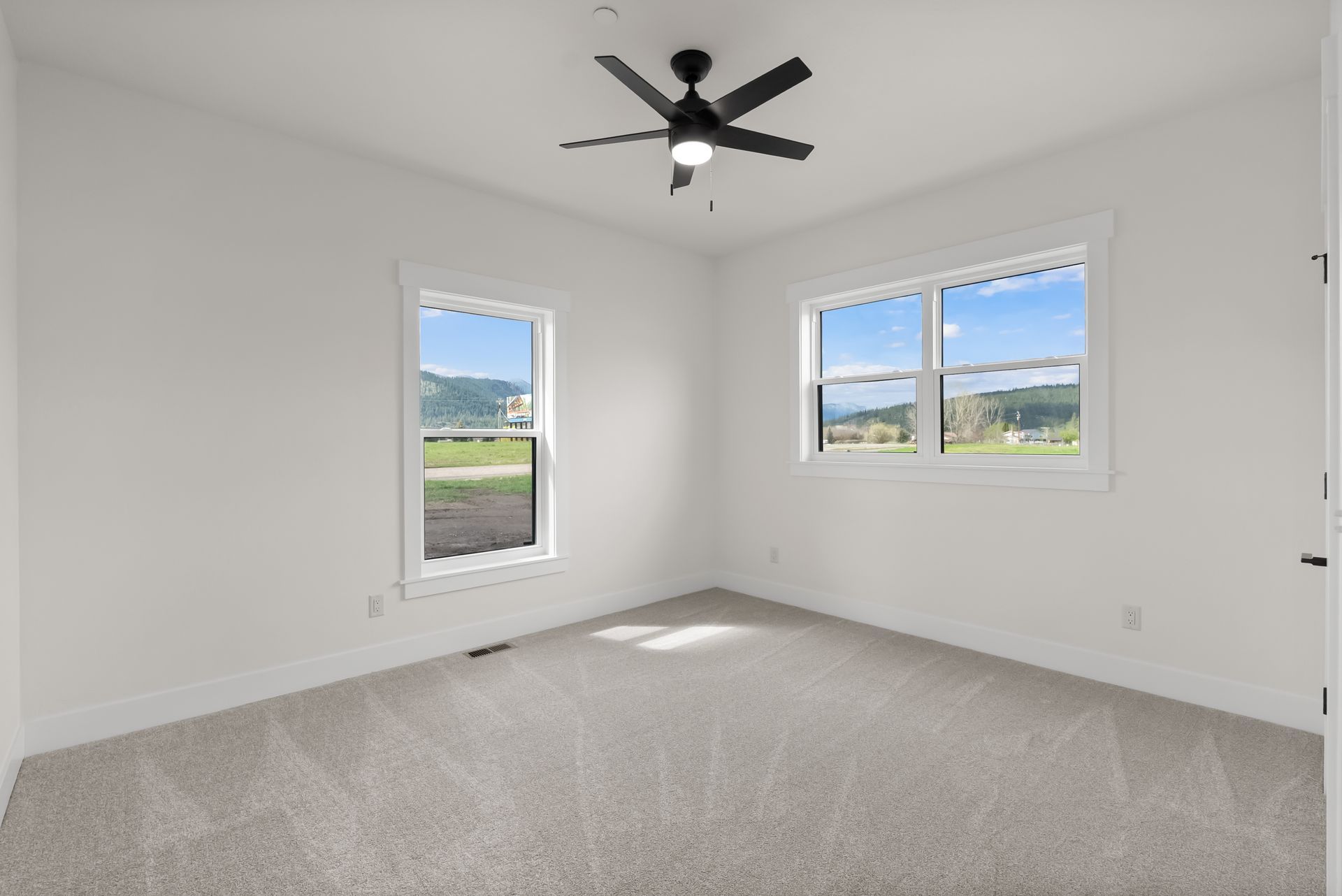 Empty white bedroom with gray carpet, black ceiling fan, and two windows overlooking a landscape.