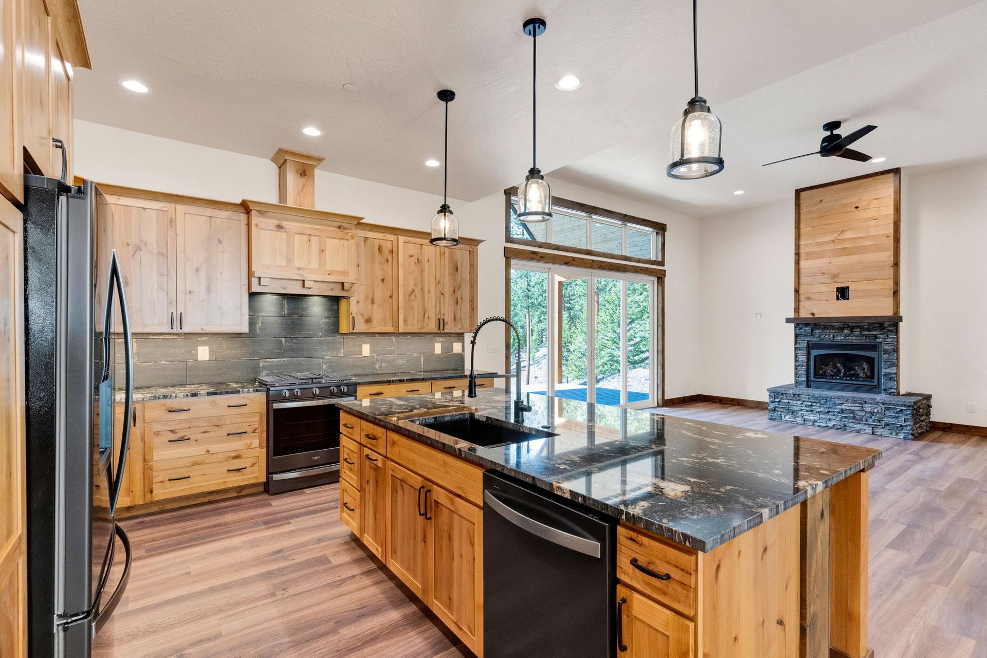 Modern kitchen with wood cabinets, black granite island, pendant lights, and a stone fireplace in the background