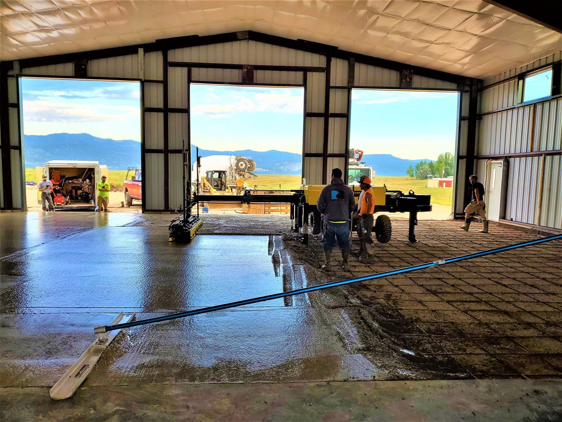 Workers smoothing wet concrete inside a large metal barn with open doors to a sunny field.