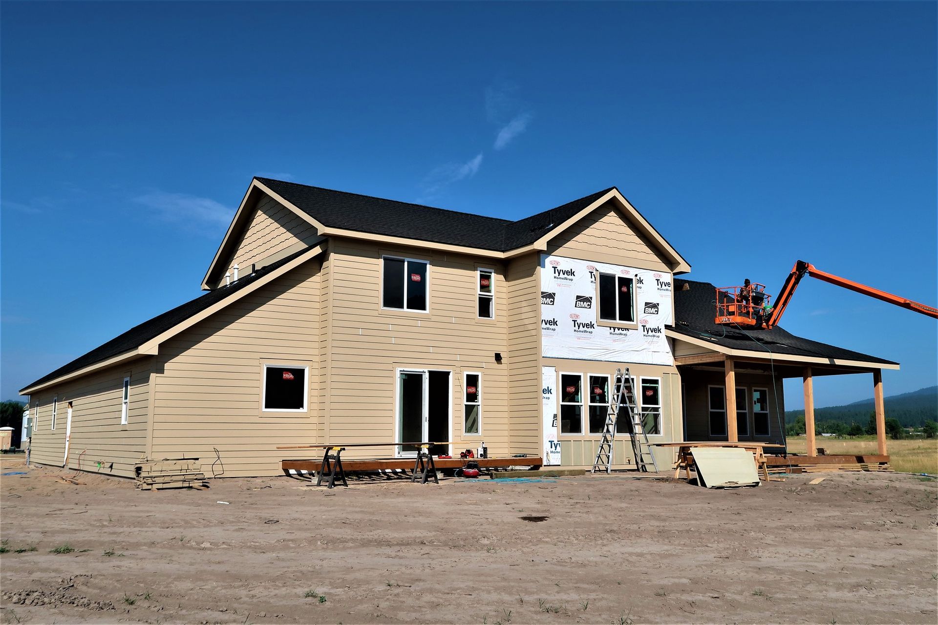 Two-story house under construction with exposed wood siding and an excavator nearby under a blue sky