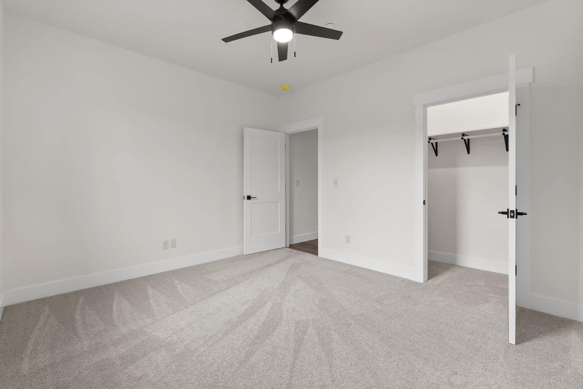 Empty white bedroom with gray carpet, ceiling fan, open closet, and open door to hallway